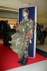 Mannequin vêtu d'un treillis militaire, d'un casque et de chaussures Rangers présentant l'équipement d'un parachutiste : un parachute EPI EPI, une gaine EL 32 et un fourreau d'arme EL 33.