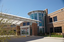 A driveway with a covered pedestrian crossing leads to automatic sliding doors with a sign above reading "Center for Advanced Medicine"; behind the entranceway, a small cylindrical glass and steel atrium leading to a brick building.