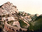 A photochorm picture of the a curving railroad bridge coming out of a tunnel bored into a large mountain. In the background another bridge and mountains are visible.