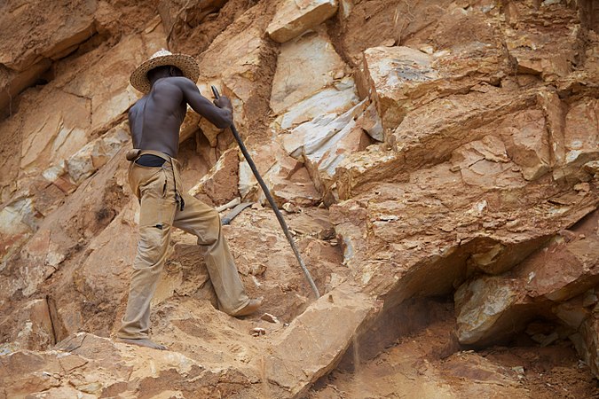 Photograph of shirtless male stoneworker in a straw hat and khaki pants testing large rocks with a staff