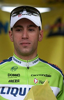 A road racing cyclist in a green and blue jersey with white trim, and a white cap with sunglasses on it.
