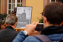 Photograph of people gathered in a town square and looking at a framed black-and-white photograph of Samuel Paty teaching, beneath which is written: "Samuel Paty, 1973–2020"