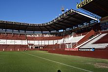 Corner of a stadium with empty maroon and white seating sections, viewed from the pitch, from which it is separated by a fence; the sky is blue and cloudless.