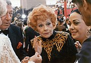 An aged Ball standing in a crowd of celebrities,  wearing a black and gold sequinned dress with her characteristic red hair, looking fragile.