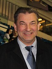 A smiling man wearing a dark suit over a light-colored shirt and blue tie. He has short and dark hair with some gray highlights.