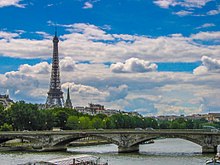 Frontale Farbfotografie einer Steinbrücke mit Verzierungen an der Vorderseite, auf dem Autos fahren. Im Hintergrund sind Baumreihen und Häuser, an deren linker Seite der Eiffelturm steht.