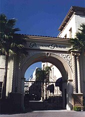 Photographie de l’arche stylisée qui marque l’entrée des studios Paramount, sous un ciel bleu, une grille fermée empêche de pénétrer dans l’enceinte, deux palmiers jouxtent l’arche.