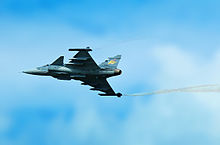 Three-quarter hind bottom view of jet aircraft in flight generating wingtip vortices, against a blue cloudy sky