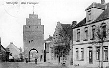 Photo en noir et blanc montrant une rue du début du XXe siècle. À l'arrière plan, on voit une tour percée d'une grande porte.