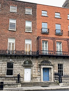 This image is of the Royal Institute of the Architects of Ireland on Merrion Square. It is a four story building with two doors next to each other one with a blue door and one with a white door. There is a silver sign that says RIAI beside the door.
