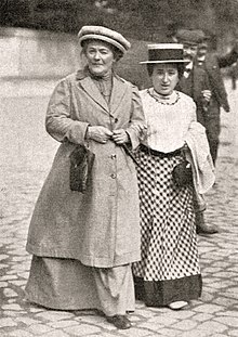 Black and white photograph of Clara Zetkin and Rosa Luxemburg. Zetkin is wearing a large overcoat and Luxemburg is wearing a checkerboard patterned skirt and a white blouse. The two are walking arms linked and are wearing hats.