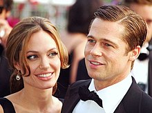 A Caucasian man and woman in the foreground of the image, while others are visible behind them. The woman has brown hair, which is tied back. The man has his dark brown hair parted. He is wearing a black suit and bow-tie with a white shirt.