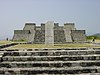 Vue générale d'une pyramide du site de Xochicalco. Au premier plan, un escalier de pierre accède à une plate-forme au centre de laquelle s'élève une stèle gravée de deux reliefs. La pyramide en elle-même est visible à l'arrière-plan.