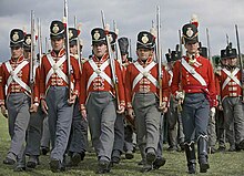 Photo of men dressed in British uniforms of the early 1800s. They wear red coats with white cross belts, gray trousers, and black shakos.