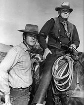 la photo noir et blanc montre à gauche Clint à pied, à côté d'un cavaler monté. Ils portent le costume des cow-boys et un lasso pend à la selle.