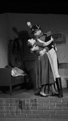 Photo prise par Fred Erismann en 1937 lors de la production de la pièce au Stadttheater Bern. Elle représente sur une scène de théâtre dans un décor représentant un pièce d'un appartement un acteur costumé en soldat prend dans ses bras une femme.