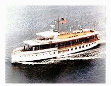 A color image of a very large yacht, in the water, with an American flag flying.