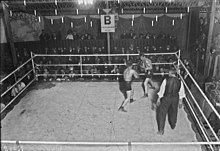 Photographie en noir et blanc de deux boxeurs s'affrontant sur un ring devant quelques rangées de spectateurs habillés en costumes.