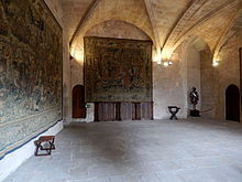 Photo couleur d'une vaste salle voutée d'arc brisé. L'appareil en pierre de taille est partiellement masqué par des tapisseries murales tissées. Les portes d'accès sont voutées en plein cintre.