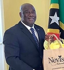 Alexis Jeffers. Ein untersetzter, glatzköpfiger Afroamerikaner im Anzug mit einer Geschenktüte von Nevis. 2022.