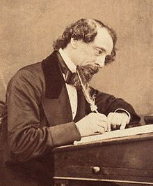 Photographie sépia de Charles Dickens écrivant sur un pupitre avec une plume.