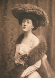 A young white woman wearing a large plumed hat, a white dress, and a fur stole.