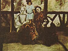 A colour-tinted monochrome photograph of a group of actors. At the rear, a young girl. In front of her, the iron man and scarecrow from 'The Wizard of Oz'; in front of them, people dressed as a lion and another animal.