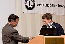 a Native American Hispanic man dressed in a brown suit shakes hands with a younger man with curly hair wearing a dark blue suit. Behind the two men is a Learn and Serve American banner.