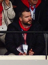 Un homme assis, vêtu d'un manteau noir et d'un foulard rouge.