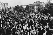 Eine Menschenmenge auf einer Straße ist im Vordergrund zu sehen, dahinter marschieren Truppen mit Gewehren. Im Hintergrund sind Gebäude der Altstadt zu sehen