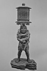 Ryūtōki. Vue de face d'une statue trapue avec un visage de démon. Il porte la lanterne en équilibre sur la tête. Il tient son poignet droit avec la main gauche. Photographie noir et blanc.