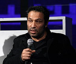 close-up of Shoja Azari wearing a black jacket, appearing to speak into a handheld microphone, looking right of camera