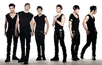 Six men wearing form-fitting black sleeveless shirts, leather pants, and combat boots. They have prominent eye makeup and each has a different gelled hairstyle.