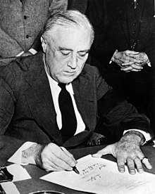 Photo noir et blanc d'un homme aux cheveux gris, en costume et arborant un brassard noir, assis à une table et qui signe un document.