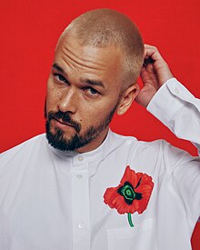 Justin Nozuka wearing a white, collarless shirt with a poppy flower over the left breast pocket, head tilted to the right with left hand scratching back of head, looking directly at camera