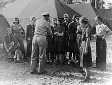 A photograph from 1945, taken outdoors, in front of a series of large tents; about 12 young white women, mostly in denim dresses, are standing and sitting; a man in uniform has his back to the camera.