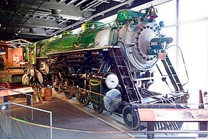 A green steam locomotive with a 4-6-2 wheel arrangement (four leading wheels, six driving wheels, and two trailing wheels) and its tender with a horse and cart in the foreground