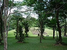 A large grassy mound seen through widely spaced trees scattered across a flat grassy foreground. A stone stairway climbs the mound and several stone monuments stand in front of this stairway. More trees form the backdrop to the mound.