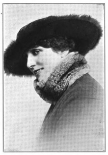 A smiling white woman wearing a dark, wide-brimmed hat, and a scarf wrapped around her jawline, obscuring her chin and nape