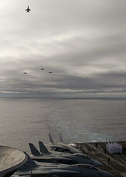 Photographed from the flight deck of an aircraft carrier: three airplanes are flying in formation while approaching, and a fourth plane is climbing up and away from the formation to signify the missing man.