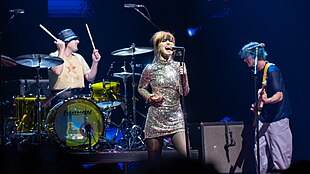 In the background, drummer Zac Farro sits in front of a yellow-tinged drum kit with his drumsticks in the air. Hayley Williams, a white woman with red, pulled-back hair, wearing a silver dress, sings into the microphone. To her right, guitarist Taylor York faces her and plays the guitar.