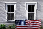 oil painting of a white clapboard house,two windows with flag draped along bottom sills, weeds along base