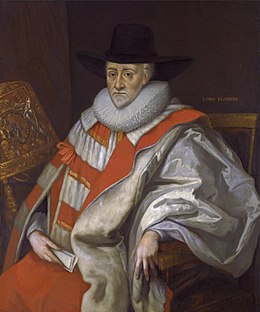 Un homme aux cheveux et à la courte barbe blanche, assis dans un fauteuil. Il est vêtu d'amples robes rouges et blanches et d'un chapeau noir à large bord.