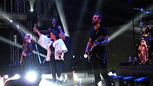 Photo en couleur d'un groupe de musique sur une scène, une chanteuse brune habillée de blanc lève un bras