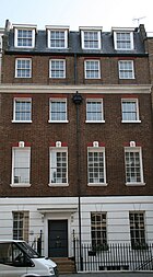 A terrace building. Its ground floor has plaster render inscribed to look like stone, the middle three are red brick, and the top is an attic. Each floor has four sash windows with a dozen or more panes each, except that the bottom floor has a door in place of the second window.