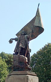 Фотография парка, в котором стоит большая статуя, изображающая бородача, стоящего на скале, одетого в длинное пальто и держащего шляпу в правой руке, а левой рукой сжимающего большое знамя.