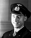 A smiling man wearing a peaked cap e dark military uniform with an Iron Cross displayed at the front of his uniform collar.