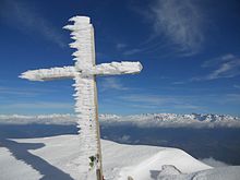 Croix sommitale en bois sur laquelle se sont formés des cristaux de glace dans le sens du vent et une longue chaîne de montagne enneigée en arrière-plan sous un ciel azur.