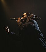 A black man, dressed in all black, stands side on to the camera. He is performing on stage with a microphone in his hand