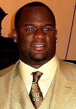 A headshot of a young black man with short hair, two earrings, a gold suit and tie.